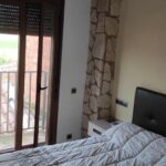 Habitació de matrimoni on es veu el llit, una porta que dona al balcó, un capçal de llit de color negre i una taula de nit blanca amb un detall de color negre.