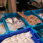 Expositor d'una peixateria on hi ha diferents productes: galeres, lluç... dintre de safates de color blau, al darrere de l'expositor hi ha una noia vestida de negre d'esquena, netejant peix.