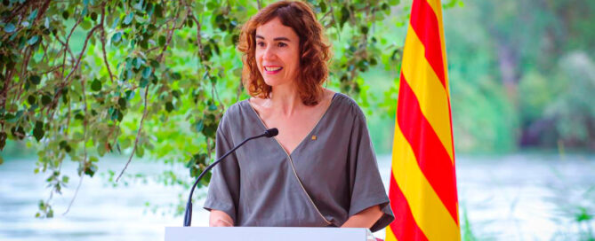 Dona fent un parlament al darrere d'un faristol gris on hi ha el logotip de la Generalitat de Catalunya i Sempre Endavant, al darrere es veu el riu i la bandera de Catalunya.