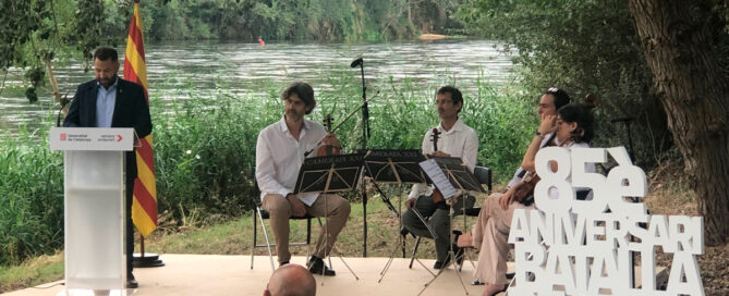 Acte de l'aniversari de la Batalla de l'Ebre al costat del riu on hi ha un home fent un parlament, al darrere hi ha una bandera i a l'altra part de l'escenari hi ha tres homes i una dona asseguts amb violins a la mà.