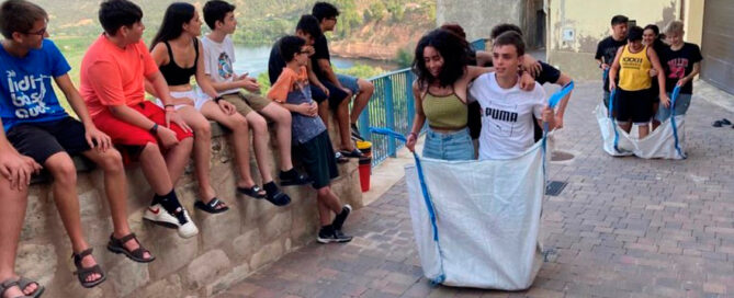 Joves del Camp de Treball jugant a la cursa dels sacs a un carrer d'Ascó amb vistes al riu Ebre.