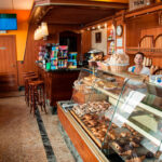 Interior d'una pastisseria i/o forn de pa on predomina el color de fusta, ja que les cadires, les taules, els tamborets, la paret... són de fusta. També hi ha un expositor amb vidriera on es veuen diferents productes de pastisseria: croissants, napolitanes...