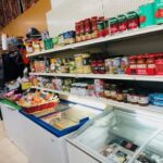 Fotografia de l'interior de la botiga on es veuen dos congeladors i una nevera amb productes dintre. A dalt d'aquestes, hi ha estanteries amb salses de tomata envasada, espàrrecs i tot un variat de productes envasats en lata.