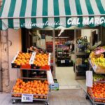 Fotografia de l'exterior de la botiga on es veuen exposades taronges i plàtans i un para-sol on hi ha escrit:"Alimentación Cal Marcos".