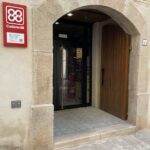 Exterior de la botiga ferreteria Biarnés, on es veu una paret de pedra amb una arcada i una porta de fusta oberta. Més endins, hi ha una porta d'alumini amb vidre.