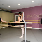 Fotografia d'unes ballarines practicant ballet amb una banca, la paret del darrere és de color rosa amb el logotip de color daurat de "Le’scènica, Escola de Dansa i Arts Escèniques".