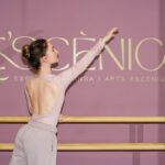 Fotografia d'una ballarina practicant ballet amb una banca, la paret del darrere és de color rosa amb el logotip de color daurat de "Le’scènica, Escola de Dansa i Arts Escèniques".