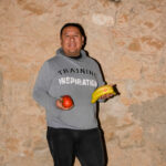 Fotografia mig pla d'un home vestit amb pantalons negres i una dessuadora de color gris amb plàtans a la mà esquerra i tomates a la mà dreta.
