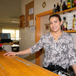Dona al darrere d'una barra d'un bar on hi ha botelles, gots, copes...